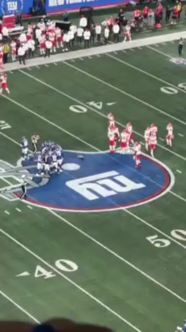 The video shows a bird's-eye view of an NFL football game between the New York Giants and the Kansas City Chiefs. The Giants are in blue and white uniforms while the Chiefs are in red and white uniforms. Both teams are gathered around the center field logo at the start of the game. There are also other people on the sidelines wearing jackets. The field is a green color with yard markers in white. The stadium has fans watching in the stands.