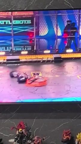 A screen displays a robot combat event. Two robots, one red and black, and another predominantly black, are engaged in a battle. The red and black robot is in motion, as is the black robot. The robots are on a grey arena floor with yellow and blue markings. In the background, a large screen displays "BATTLEBOTS" and blurry figures. The video cuts to the red and black robot flipping over and then briefly showing the black robot as it moves. The battle continues with the robots colliding. The black robot appears to be throwing the red and black robot. The red and black robot spins violently before being impacted. The black robot then spins the red and black robot again. The robots are seen in motion on the arena floor. The black robot then appears to strike the red and black robot, causing it to flip upside down. The robots continue to move around the arena.
