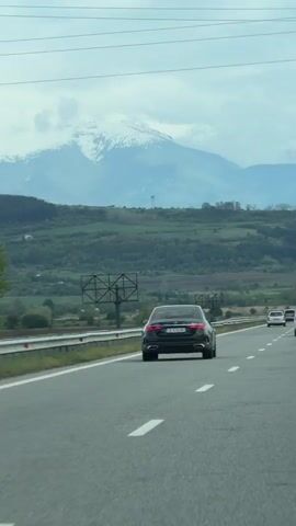 The video, shot from a moving vehicle, depicts a highway scene with a prominent mountain range in the background. The foreground consists of a multi-lane highway with vehicles traveling on it. A dark-colored sedan is directly in front of the camera car, and other cars are visible in the distance. The road is asphalt, and white lane markings are present. A metal barrier runs along the side of the highway. Beyond the barrier, there is a green landscape of rolling hills and fields. In the distance, a mountain range dominates the horizon. The peaks of the mountains are covered in snow. The sky is overcast with gray clouds.