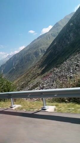 A wide shot captures a road traveling alongside a steep mountain slope under a clear blue sky with a few wispy clouds. The road, paved with asphalt, is in the foreground and occupies the bottom third of the frame. A metal guardrail, silver in color with vertical supports, runs along the edge of the road, separating it from the mountain. The mountain itself is rugged and covered in a mix of green vegetation and rocky scree. Patches of pinkish-purple flowers are visible scattered on the lower slopes. The sun casts shadows from the guardrail onto the road surface, indicating it is daytime. The perspective suggests the camera is moving, likely from within a vehicle.