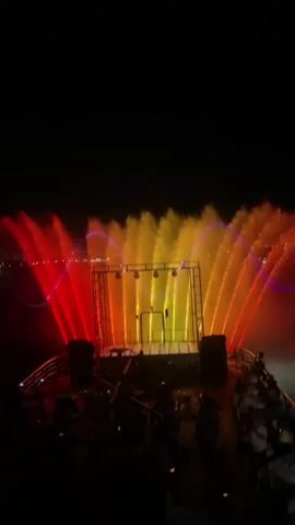The video shows a nighttime outdoor water and light show. In the foreground, a crowd of silhouetted people are gathered on a dark platform, likely a boat deck, facing a stage. The stage is rectangular and illuminated with various colored lights, primarily red and yellow. Behind the stage, a large water fountain display is in motion, with jets of water shooting up and being illuminated by colorful lights. The lights on the water change colors, cycling through red, yellow, and green, with some blue light effects also visible. The background is dark, with a faint outline of distant lights suggesting a city or shoreline. The water jets create a dynamic visual effect, appearing to dance and sway in sync with the changing lights.