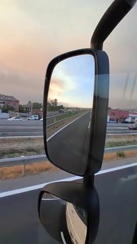 The video shows a view from a moving vehicle, looking at the road ahead and the sky. In the foreground, a large side mirror of a truck is visible, reflecting the scene. The road appears to be a highway with multiple lanes and a guardrail on the side. The sky is filled with clouds and a bright sun, suggesting it is either sunrise or sunset, casting a warm orange and yellow glow. Along the side of the road, there are some buildings and trees. The camera pans slightly, showing the ongoing movement of the vehicle and the changing scenery. There are no people visible in the video. The environment appears to be an urban or semi-urban area.