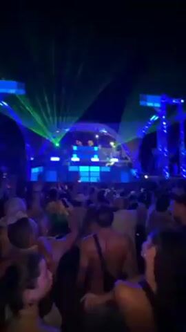 A crowd of people are attending an outdoor music festival at night. The stage is lit with blue lights and has an intricate geometric design. There are also green lasers shining over the crowd. Some people are raising their hands in the air and dancing. The environment appears to be lively and energetic.