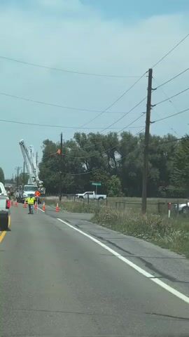 The media file depicts a scene of road work with utility maintenance taking place on a rural road. The foreground shows a paved road with yellow double lines, indicating a two-way traffic flow. A white pickup truck is visible in the left lane, and orange traffic cones are strategically placed along the road to delineate the work area and guide traffic.

In the middle ground, several utility trucks, including a large bucket truck with its arm extended, are parked on the side of the road. Workers in high-visibility vests are present, one of whom is holding a "SLOW" sign, indicating that traffic is being managed. Power lines and utility poles are visible overhead, suggesting that the work involves power line maintenance or repair.

The background features a line of trees and vegetation, typical of a rural or semi-rural setting. A fence runs along the right side of the road, bordering a grassy field. A street sign reading "Arlington Dr" is visible on the right, providing a specific location cue. The sky is partly cloudy, suggesting it is daytime. There are no overt emotional expressions captured, but the presence of road work and traffic control implies a disruption or planned activity. The overall scene conveys a sense of ongoing utility operations.