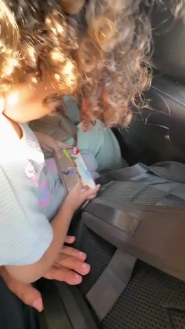 The video begins inside a vehicle, showing the curly brown hair of a child in the foreground. The child is holding a smartphone displaying a game. Another child, wearing a light blue shirt, is visible behind the first child, partially obscured by a large black bag with straps. The scene transitions to the exterior of the vehicle, showing a paved road under a bright sunny sky with scattered clouds. A person's arm, wearing a light-colored sleeve, is visible on the left side of the frame, possibly holding the camera. The road appears to be in a rural or desert environment with sandy terrain and vegetation on the right side of the road. The sun is shining brightly, creating a glare on the road.