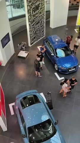 The video shows an overhead view of an Audi dealership. Several people are present, looking at various Audi car models. One blue Audi hatchback is in the center of the frame with a group of people around it. To the left, a light blue Audi coupe is visible. To the right, a blue Audi hatchback and a silver race car are displayed. In the background, a white concept car is also on display, with a few people gathered around it. The dealership has a modern interior with tiled floors and bright lighting. Various posters and displays are visible on the walls.