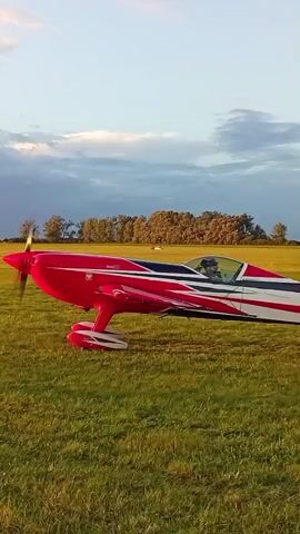 A red, white, and blue stunt plane is parked on a grassy field. The plane has a single engine and a two-person cockpit. The pilot is visible in the cockpit, wearing a helmet. The propeller is spinning, indicating the engine is running. The sky is partly cloudy, with a mix of blue sky and darker clouds. Trees are visible in the background. The sun is low in the sky, casting a warm light on the scene. The video is taken from a low angle, looking up at the plane.