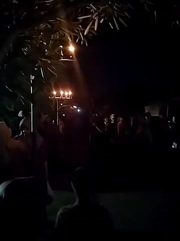 A nighttime outdoor scene illuminated by a single streetlight and a set of candles on a pole. A crowd of people are gathered, many wearing headscarves. The lighting is dim and casts long shadows. The overall atmosphere appears to be one of a gathering or event.