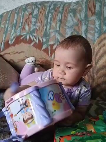 A baby lies on its stomach on a couch, playing with a toy drum. The baby is wearing a purple onesie and socks. The drum is pink and white with colorful illustrations. The baby puts the drum in its mouth. The couch has a patterned fabric. To the right of the drum is a green fabric book with animal illustrations. The video is approximately 4 seconds long.