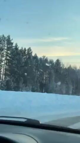 The video shows a view from inside a car driving along a road. On the right side of the road is a thick layer of snow, adjacent to a forest of frosted evergreen trees. The sky is light blue with hints of peach and yellow at the horizon. The windshield wipers are visible in the lower part of the frame. Music is playing in the background.