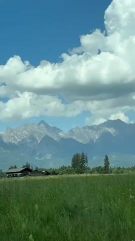 The video shows a rural landscape with a bright blue sky and fluffy white clouds. In the foreground, a vast green field stretches across the frame. In the middle ground, a cluster of trees stands to the right of a house with a dark roof and light-colored walls. Further in the background, a mountain range with rugged peaks and some snow-capped tops dominates the horizon. A small wooden building, possibly a shed, is visible on the left side of the frame. Two circular traffic signs are mounted on a pole on the left side of the frame. The scene appears to be a sunny day in a mountainous region.