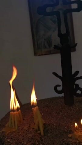 THE VIDEO SHOWS A CLOSE-UP OF SEVERAL LIT CANDLES ON A SANDY SURFACE. IN THE FOREGROUND, TWO CANDLES ARE BURNING BRIGHTLY, WITH FLAMES DANCING. TO THE RIGHT, ANOTHER CANDLE IS VISIBLE, ALSO LIT. BEHIND THE CANDLES, A SILHOUETTE OF AN ORNATE CROSS IS SEEN, PARTIALLY OBSCURED BY SHADOWS. ABOVE THE CROSS AND TO THE LEFT, A FAINT IMAGE OF WHAT APPEARS TO BE A RELIGIOUS ICON OR PAINTING IS VISIBLE ON A PLAIN WALL. THE LIGHTING IS DIM, SUGGESTING AN INDOOR SETTING, POSSIBLY A CHURCH OR CHAPEL. THE AMBIANCE IS PEACEFUL AND SOLEMN.