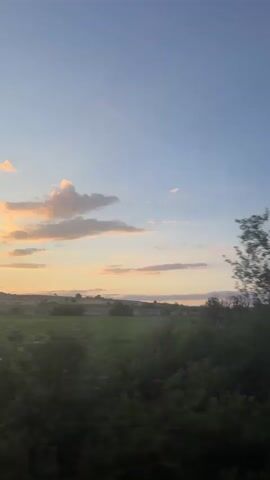 The video displays a continuous view from a moving vehicle, showcasing an outdoor landscape. The sky occupies the upper two-thirds of the frame, transitioning from a light blue at the zenith to warmer shades of yellow, orange, and pink near the horizon, indicative of twilight. Scattered cumulus clouds, some illuminated by the warm light, are visible across the sky. The lower third of the frame consists of a blurred expanse of green fields and dark green foliage, primarily trees and bushes, suggesting a rural environment. The motion blur indicates rapid horizontal movement relative to the stationary landscape. At approximately 0:02, the frame briefly shows reflections of the sky and clouds on the vehicle's windows as it passes close to dense, dark green trees before returning to the open landscape view.