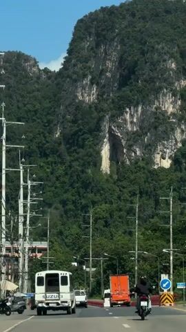 The video shows a view from a moving vehicle on a road. A large, green, mountainous cliff face with a cave opening is visible in the background. In the foreground, there are several vehicles on the road, including a white van with "SUZUKI" on the back, an orange truck, and motorcycles. Utility poles with wires line the side of the road. The sky is blue and clear.