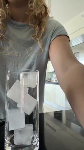 A person with curly blonde hair, wearing a light green t-shirt, is preparing a drink in a kitchen. They pour green powder from a pink and green labeled packet into a tall clear glass filled with ice cubes. The kitchen features a black countertop, stainless steel appliances, and light-colored cabinets. A red bracelet is visible on the person's left wrist.