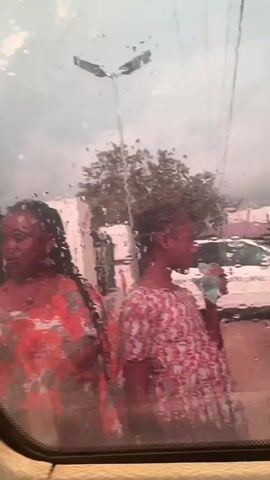 The video is a close-up shot through a rain-streaked window. Two people are visible outside. On the left, a person with dark hair and braids is wearing a red and white patterned top. They are looking to the left. To their right, another person with dark hair and braids, possibly a young woman, is wearing a pink and white floral top. She is holding a bottle to her lips and appears to be drinking. In the background, there are cars, trees, and buildings, suggesting an outdoor setting, possibly a street or parking lot. The overall atmosphere is muted due to the rain and the window's condition.