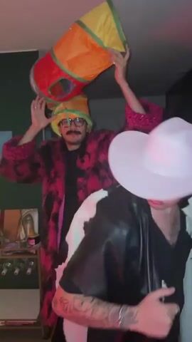 The video shows a person wearing a pink and black fur coat and glasses, holding a colorful tunnel toy over their head. The person appears to be dancing at a party, as two other individuals are seen in the background, one wearing a black leather jacket with a cow print, and another with a cowboy hat. The room is dimly lit, with what looks like a wine rack in the background.