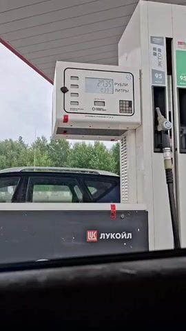 This is a video taken from inside a car at a Lukoil gas station. The camera is focused on a gas pump display that shows the rising cost and liters of fuel being dispensed into a car. The car appears to be a dark-colored SUV. The background consists of trees and a cloudy sky. The gas pump displays the brand name "Lukoil" and the fuel type "95".