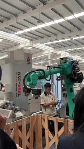 The video shows a light and spacious factory environment with high ceilings and abundant overhead lighting. Focus is on a man standing behind a wooden barrier, operating a green robotic arm connected to a CNC Vertical Machining Center. The machine, labeled "BSC-850L", has its name and the company logo, "SHENCHENG", prominently displayed. The man is focused on a handheld control panel, presumably programming or adjusting the robot's movements. People can be seen watching him from behind the wooden barrier. The scene suggests a demonstration or training session highlighting automated manufacturing processes. The setting is clean and organized, indicating a modern industrial facility.