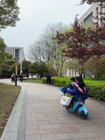The video shows two people on a light blue electric scooter driving on a paved walkway. The person in the back has long dark hair and is wearing a blue jacket and pink strap. The person driving the scooter is also wearing a blue jacket. They are carrying a white shopping bag. The walkway is lined with green bushes and trees. In the background, there are buildings, including one with a grid pattern. There are a few people walking and riding bicycles in the distance. The weather appears overcast.