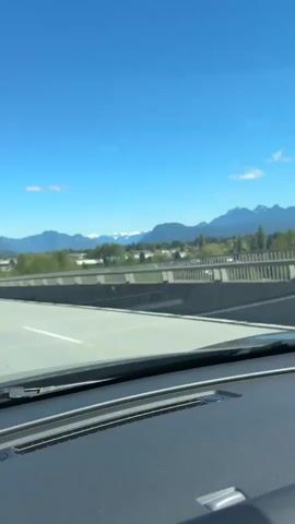 A car drives on a highway overpass with mountains visible in the distance under a clear blue sky with a few clouds. The camera is positioned behind the car's dashboard, capturing the view through the windshield. The overpass has gray concrete barriers and metal railings. Trees and buildings are visible between the overpass and the mountain range. The driver appears to be moving at a consistent pace. A tall streetlight is visible on the right side of the road.