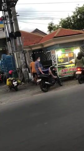 The video shows a street scene with several motorcycles and people. In the foreground, there is a road. To the right, there is a food stall with a sign that reads "SINGKONG KEJU". A woman wearing a face mask is sitting on a motorcycle in front of the stall. Behind her, another person is standing. To the left, there is a utility pole and several other motorcycles, one of which has a red helmet. The background shows a building with a tiled roof and trees. The overall lighting suggests it is daytime, possibly overcast.