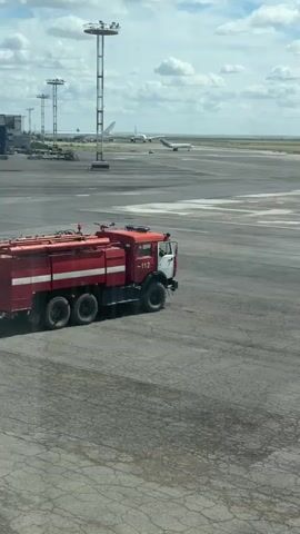The video shows a view of an airport tarmac under a cloudy sky. In the foreground, a red fire truck is parked. In the background, several airplanes are visible on the runway, and tall lights stand along the runway. The tarmac appears to be made of cracked asphalt.