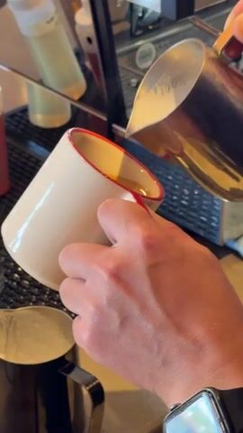 A person's hands are visible in the frame, holding a white ceramic mug with a red rim and handle. The mug contains a golden-brown liquid, likely coffee, with some crema on top. The person is pouring steamed milk from a silver metal pitcher into the mug. The milk is creating a swirling pattern on the surface of the coffee, indicating latte art is being created. The background is blurred but appears to be a cafe or coffee shop environment, with a coffee machine and other containers visible. The lighting is bright, suggesting indoor lighting.