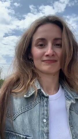 The video shows a woman smiling at the camera, standing on a paved path outdoors. She has long, wavy brown hair, is wearing a white t-shirt and a light blue denim jacket. She has hoop earrings and a small piercing on her nose. Behind her, there are stairs leading up a grassy hill and stone steps. There are also several dome-shaped structures with brown roofs, resembling covered seating areas, in the background. The sky is partly cloudy. The woman is looking directly at the camera, and seems to be enjoying the sunny day.