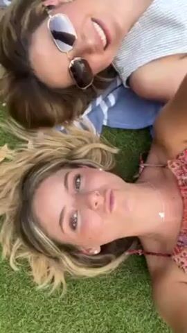 Here is a concise description of the video:

The video shows two women lying on the grass, posing for a selfie. One woman is wearing sunglasses and a striped shirt and is on a striped towel. The other is wearing a pink dress and a necklace with a small pendant. They are both smiling at the camera. The background is green grass. The camera is oriented at a downwards angle towards the women.