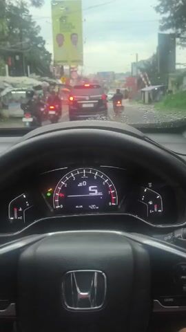 A driver navigates through rainy street traffic at approximately 6 kilometers per hour. The viewpoint is from inside the car. A large advertisement for the Golkar political party is visible next to the road. Other cars and motorbikes are present on the road.