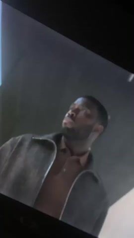 The video displays a television screen showing a man, likely in a scene from a movie or show. The man, with dark skin and a short beard, is wearing a dark jacket over a brown polo shirt and is looking upwards with a contemplative expression. Subtitles indicating singing, "♫ Ahh, ahh, ahh, ahh, ahh, ahh ♫", are visible on the screen. The camera then pans away from the screen, revealing a modern interior room with large windows. Through the windows, a bright, sunlit landscape can be seen, suggesting it is daytime. The overall location context is Gosa, Nigeria.