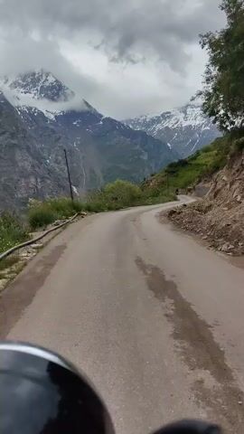 The video shows a winding paved road in a mountainous region. The road is wet, suggesting recent rain or mist. The weather is overcast, with dramatic clouds hanging over snow-capped mountains in the distance. The mountains are rugged and imposing, with green slopes leading up to the rocky, snow-covered peaks. Along the side of the road, there are some retaining walls made of stone, and loose rocks are visible on the embankment. There is some vegetation, including grass and bushes, growing on the hillsides.

A few small structures, possibly houses or guesthouses, are visible on the green slopes higher up the mountain. A person on a motorcycle or scooter can be seen in the distance, traveling on the road. The perspective suggests the video is being filmed from the point of view of someone riding a motorcycle or scooter. The foreground shows a portion of the handlebars or helmet of the rider.

The scene evokes a sense of travel and adventure, with the dramatic mountain landscape and the winding road suggesting a journey through a remote and beautiful area. The overcast weather adds to the dramatic and slightly moody atmosphere. The location is identified as Gemur, India, which is known for its scenic mountainous terrain. There is no visible text in the video.