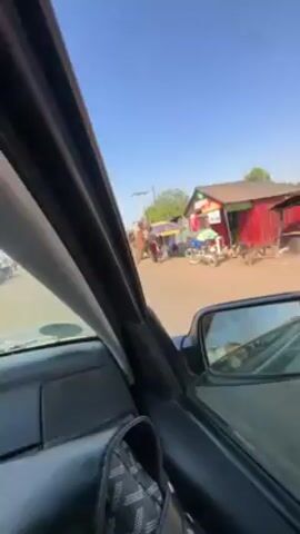 The video, taken from inside a car, shows a street scene in what appears to be a less developed area. The car is stopped in traffic on a dirt road, with simple structures and makeshift shops lining the street. People are walking around, and other vehicles are visible. The sky is clear and blue.