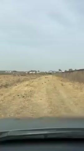 The video shows a car driving on a dirt road through a dry, grassy field. In the distance, there are several houses. The sky is overcast. A low wall runs along the right side of the road. The video is taken from the perspective of the driver.