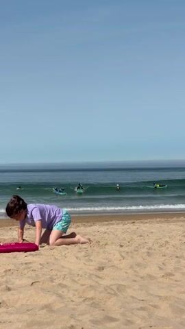 The video opens with a young person on their hands and knees on a sandy beach, facing the ocean. Their left hand is on a pink object. In the middle ground, two surfers are riding waves on surfboards. The surfer on the left is in front and slightly to the left of the surfer on the right. Both surfers are wearing wetsuits with yellow and blue accents. The ocean is calm with gentle waves breaking on the shore. The sky is clear and blue. The scene is a beach on a sunny day.