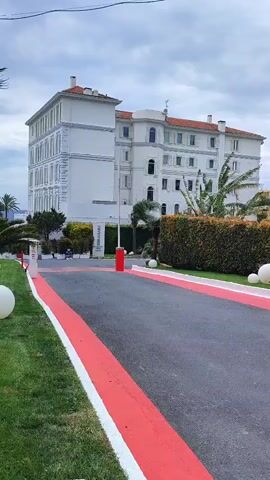 The camera films a large white hotel from an exterior location. The hotel has multiple windows, a brown roof, and is surrounded by greenery, including shrubs and palm trees. In front of the hotel, there is a gray paved road with red and white painted stripes along its edges.  A red pole-like barrier is visible in the middle of the driveway. The sky is overcast and cloudy. The camera appears to be held by someone who is either walking or standing still. A passing motorcycle is audible in the background.