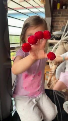 A young girl with light brown hair and a pink t-shirt with a cat design kneels in front of a black table. She holds her left hand up, with five red, spiky berries placed on her fingers. She brings her hand closer to her face, inspecting the berries. In the background, another young girl with dark hair sits in a hanging chair, wearing a white t-shirt and pink shorts. She appears to be looking at the first girl. A plate with two cherries and one raspberry sits on the table in front of the first girl. The scene takes place on a covered patio, with a brick fireplace and a suspended swing visible.