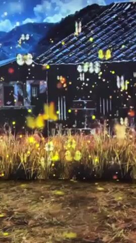 The video displays an animated scene of a house set against a night sky and mountains, with digitally-rendered wheat or tall grass in the foreground. Small, glowing yellow objects appear to float across the screen, and there's an illusion of falling snow or particles. The overall aesthetic has a dreamlike quality due to the surreal lighting and effects.