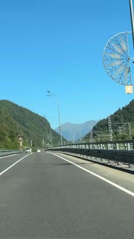 The video captures a view from inside a vehicle driving on a highway. The highway is bordered by a metal guardrail on the right and a steep, tree-covered mountainside on the left. The sky is clear and blue, with distant mountains visible in the background. Several streetlights are positioned along the highway. A few cars are seen on the road ahead.