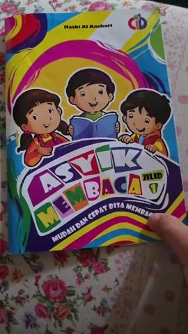 The video begins with a close-up shot of the cover of a children's book titled "Asyik Membaca Jilid 1" (Fun Reading Volume 1). The cover features cartoon illustrations of three children, two boys and one girl, smiling and looking at the book. The title and subtitle, "Mudah Dan Cepat Bisa Membaca" (Easy and Fast to Read), are prominently displayed in colorful letters. The background of the cover has a vibrant, swirly design. The book is placed on a floral-patterned fabric, possibly a tablecloth or bedding.

The camera then moves to reveal the inside pages of the book. The first page shown is the preface, titled "Kata Pengantar" (Foreword). The text of the foreword is visible, discussing the purpose and methodology of the book for teaching children to read. The author's name, "Hasbi Al Anshari," is visible at the top of the cover. The video then continues to show the pages of the book, with a finger occasionally touching or pointing to the text. The focus remains on the open book and its content. The floral fabric background is consistent throughout the video.