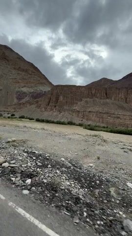 The video shows a dry, rocky landscape with mountains in the background. The sky is cloudy and grey. There is a road in the foreground and some sparse vegetation. The camera appears to be moving, possibly from inside a vehicle.