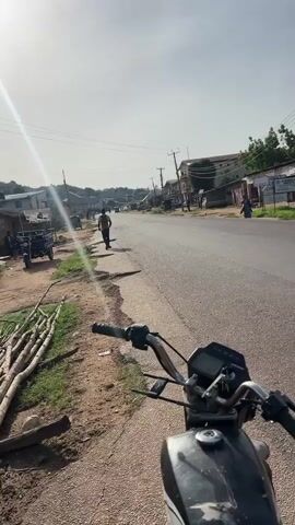 The media file shows a view from a motorcycle parked on the side of a road. The road is paved and appears to be in a somewhat rural or developing area, with buildings and vegetation lining its sides. The sun is high in the sky, indicating it is likely midday, and the weather appears to be clear and bright, with a slight haze.

Several people are visible in the scene. A person is walking down the sidewalk on the left side of the road. Further down the road, a few more figures can be seen. On the right side of the road, a small green truck is driving by, carrying several passengers in its bed.

Objects present include the motorcycle the video is presumably filmed from, a small cart with a single wheel on the left side of the road, and a pile of logs or branches also on the left. Utility poles and wires are visible overhead, suggesting infrastructure in the area. The buildings lining the road are a mix of single and multi-story structures, some appearing to be shops or residences.

The overall impression is a snapshot of daily life on a street in Rukuba Road, Nigeria. The scene is active with movement from pedestrians and vehicles, but it does not depict any specific event or heightened emotion. The lighting suggests a warm, sunny day.