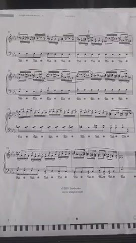 The video shows a close-up, top-down view of a sheet of music. The sheet music is on a white surface and is illuminated by overhead lighting. The music is printed in black ink on white paper and appears to be for a piano piece. The title of the piece is visible at the top of the page: "ALLEGRO CON SPIRITO" and "NUMERO 5." The music is organized into measures with treble and bass clefs. There are several lines of music visible, with measure numbers 17, 19, 21, and 23 clearly marked. The measures contain notes, rests, and dynamic markings. Below the music staff, there are pedal markings indicated by "Ped." followed by an asterisk. The bottom of the image shows a portion of a keyboard, suggesting the sheet music is intended for a pianist. There is a copyright notice at the bottom center of the page: "2023 Tombooks www.tomplay.com". The video is comprised of a series of still frames that are slightly offset from each other, creating a subtle movement effect. No people or other objects are visible in the frame. The image is slightly blurred and has a paper texture.
