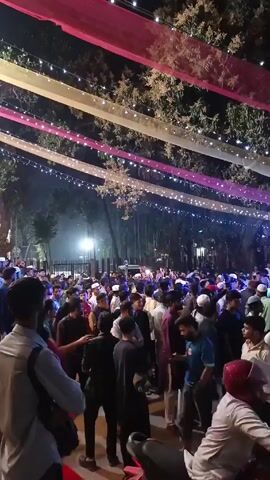 The video shows a large crowd of people gathered outdoors at night, possibly for an event or festival. Many of the individuals appear to be men, and some are wearing Muslim caps or head coverings. The area is decorated with pink and white fabric streamers and strings of small lights, creating a festive atmosphere.  There are a few motorcycles parked in the foreground. The people are generally standing and milling about, and some are looking at their phones.  The overall impression is of a lively, crowded gathering at night.