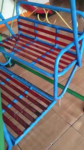 A blue metal swing set with red slats on the seats is shown. The swing set is outdoors on a tiled floor, possibly a patio or porch. The background shows part of a building and a green pole supporting the swing set. There are no people or animals visible in the video.