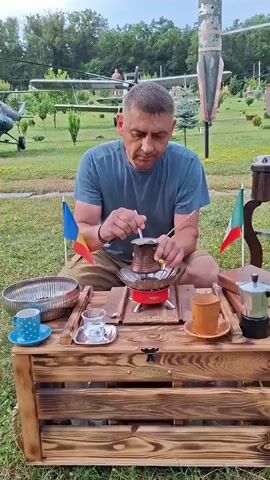 A man is preparing coffee outdoors. He is seated in front of a wooden crate which serves as a table. On the crate, there are various coffee-making accessories, including a cezve (Turkish coffee pot), a small stove, a moka pot, and a teacup with a saucer. Two small flags, one Romanian and one Italian, are placed on either side of the cezve. The man is wearing a blue t-shirt and shorts, and he appears to be stirring the coffee in the cezve. In the background, there is a grassy field with trees and what appears to be a small airplane or helicopter. The lighting suggests it is daytime, possibly overcast.