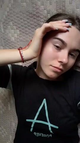 The video shows a young woman lying on her back on a textured surface, possibly a bed. She is wearing a black t-shirt with a turquoise triangular design on the left side and turquoise stripes on the sleeves. She has a red bracelet on her left wrist. The woman is looking towards the camera, with her left hand resting on her forehead. Her eyes are mostly open, and she blinks several times throughout the short clip. The lighting appears to be natural or soft indoor lighting, illuminating her face and upper body. The camera is positioned above and slightly to the side of the woman, capturing a close-up view of her face and the immediate surroundings. The video is shaky and seems to have been recorded from a handheld device.