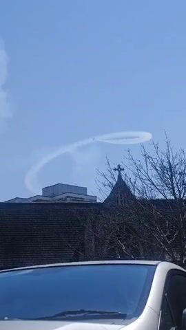 The video shows an airplane performing aerobatics in a clear blue sky, leaving a white smoke trail in its wake. The plane is seen making a large loop. Below the plane, there is a building with a dark tiled roof, a church steeple with a cross, and the top of a car in the foreground, obscuring the lower part of the scene. The presence of bare tree branches suggests it could be late winter or early spring. The overall mood appears to be bright and perhaps exciting, given the aerial display. There is no visible text in the media.