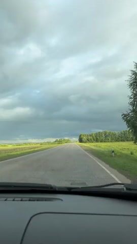 The video depicts a view from inside a car driving down a rural road. The road is paved and stretches straight ahead, with green fields on either side. A line of trees runs along the right side of the road in the distance. The sky is overcast with heavy, grey clouds. The dashboard of the car is visible at the bottom of the frame. The camera appears to be static, capturing the forward motion of the vehicle. There is no visible traffic.